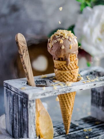 A hand sprinkling coconut chips over a Coconut Chocolate Dairy-Free Ice Cream cone placed in an ice cream stand and a wooden spoon besides it