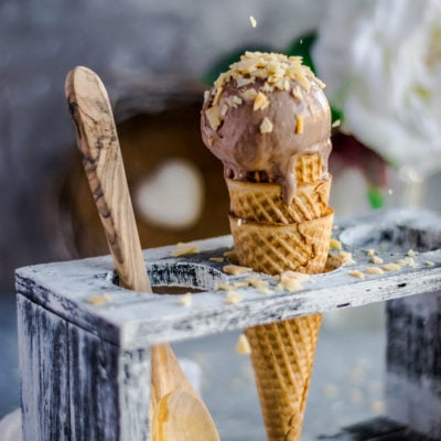 A hand sprinkling coconut chips over a Coconut Chocolate Dairy-Free Ice Cream cone placed in an ice cream stand and a wooden spoon besides it