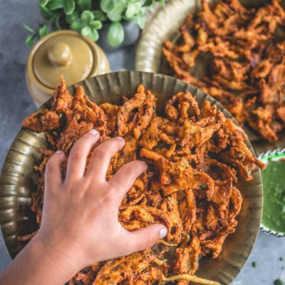 a child's hand reaching out to a plate filled with Vegan Crispy Onion Pakoda | Fried Kanda Bhajiya