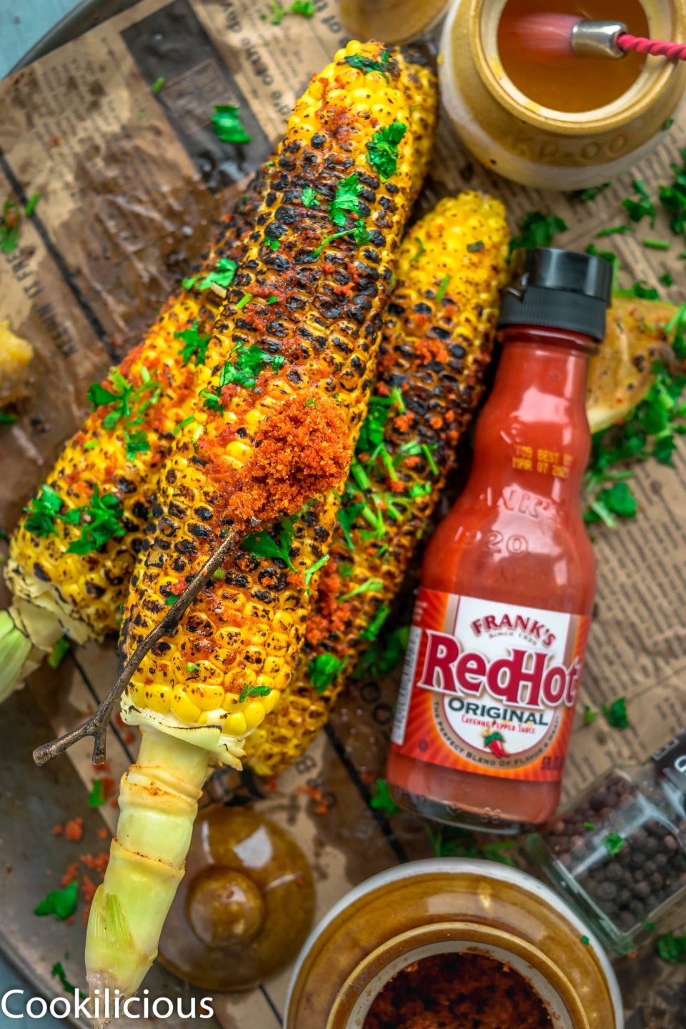 Indian Street Style Corn On The Cob on a platter with a bottle of hot sauce on the side and a spoonful of spice rub over it