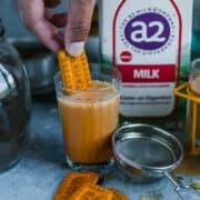 a hand dipping a biscuit into a glass of masala chai with a2 milk in the background