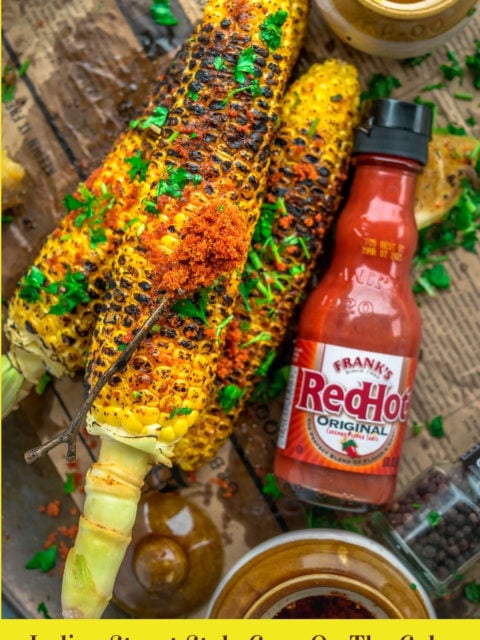 Indian Street Style Corn On The Cob on a platter with a bottle of hot sauce on the side and a spoonful of spice rub over it and text at the bottom