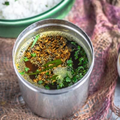 South Indian Tomato Rasam With Pink Lentils in a stainless steel vessel with a bowl of rice next to it