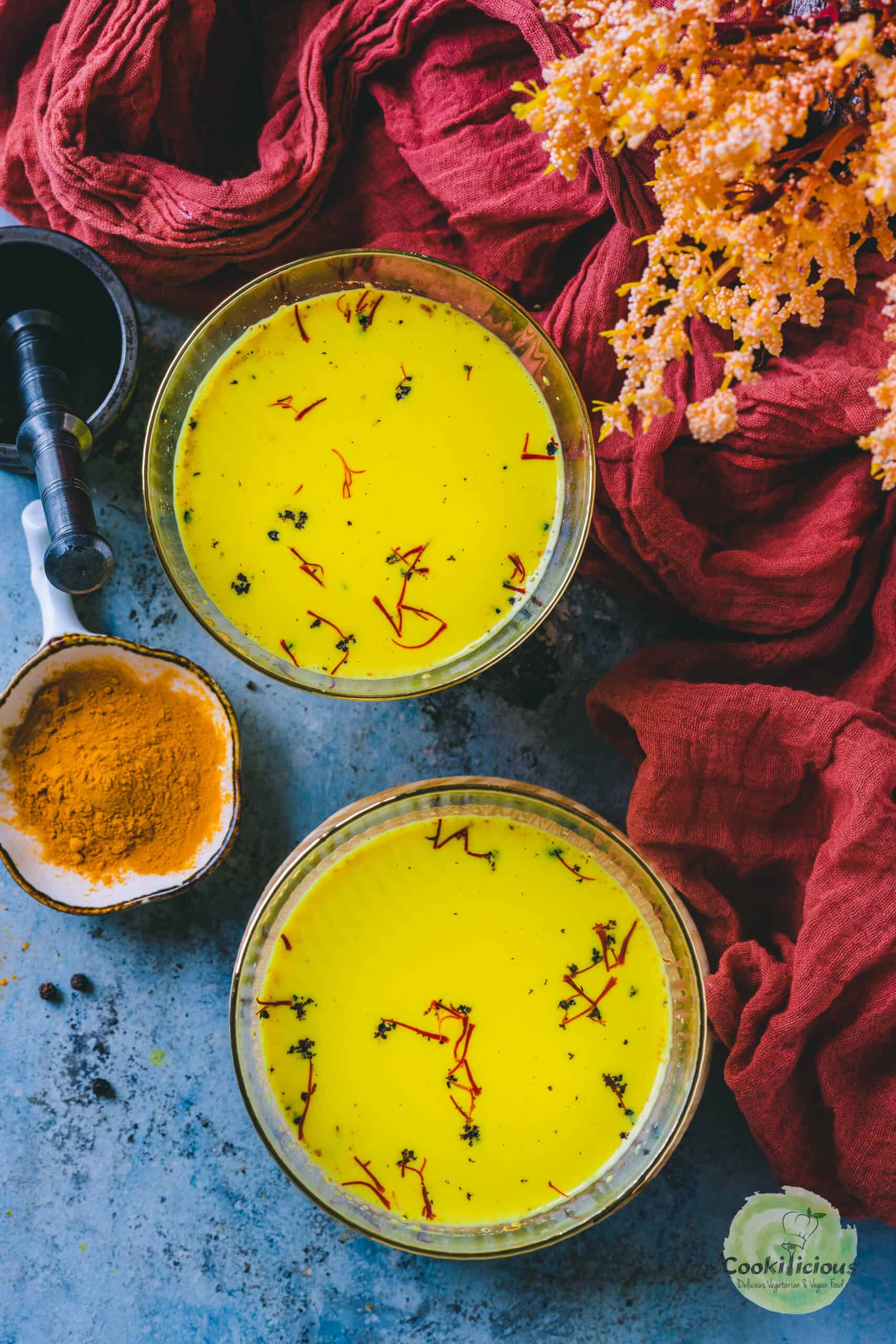 Overhead shot of two glasses of golden milk latte garnished with saffron and black pepper, with turmeric powder, and a mortar and pestle next to it.