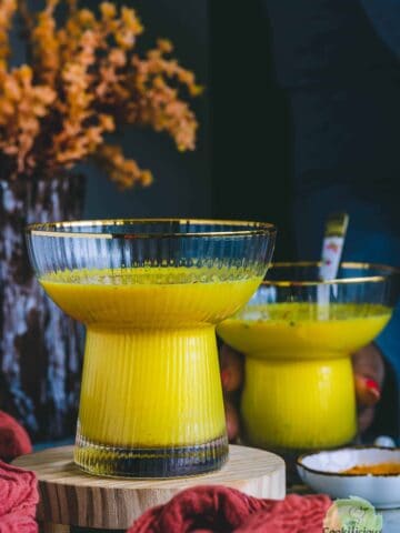 Golden milk served in two ribbed glass cups with gold rims on a wooden stand, with turmeric powder in a bowl nearby.