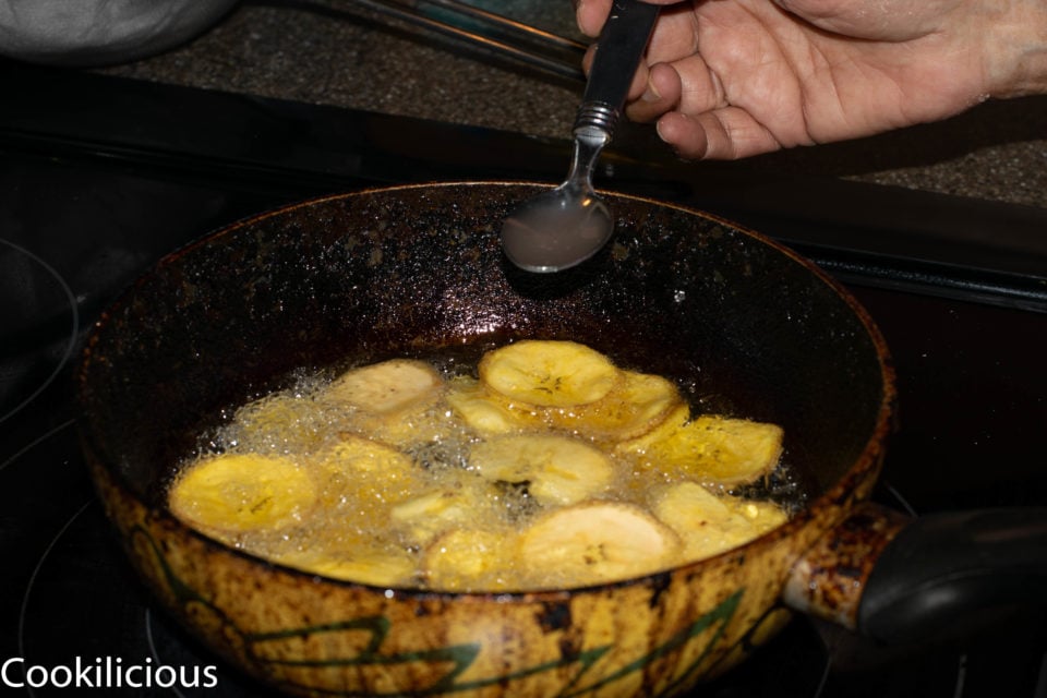 a hand adding salt water to the Fried Raw Banana Vegan Chips while they are being fried in oil
