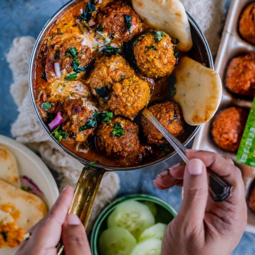 one hand holding the pan filled with Spicy Kofta Curry with Plant-Based Meatballs and the other digging in with a spoon
