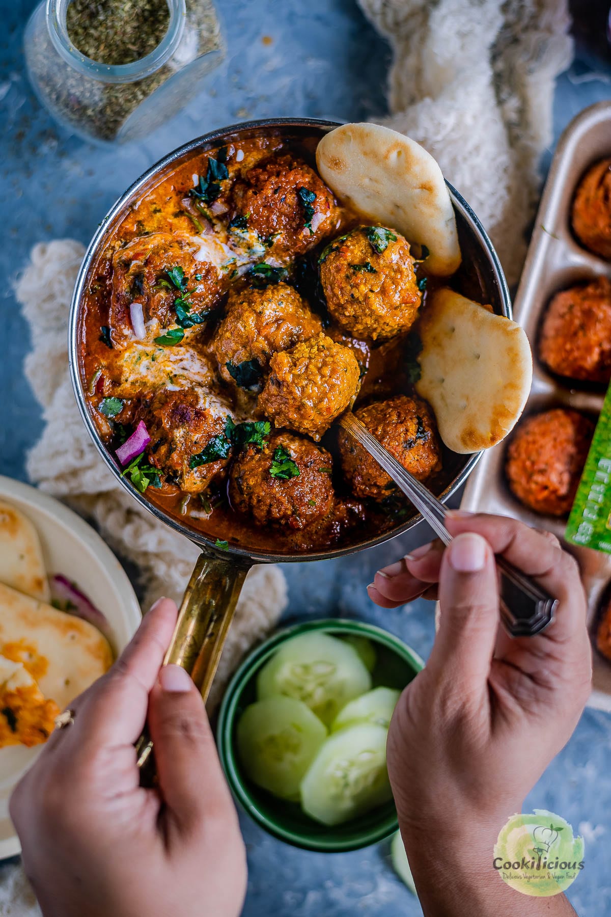 one hand holding the pan filled with Spicy Kofta Curry with Plant-Based Meatballs and the other digging in with a spoon