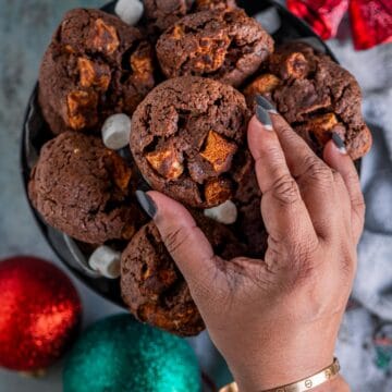 a hand picking up one chocolate chip and marshmallow cookie from the box.