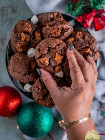 a hand picking up one chocolate chip and marshmallow cookie from the box.