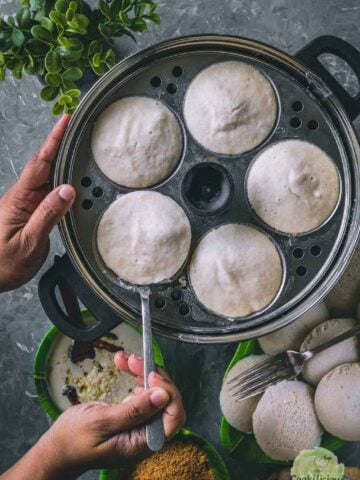 a pair of hands removing idlis from the cooker using a spoon