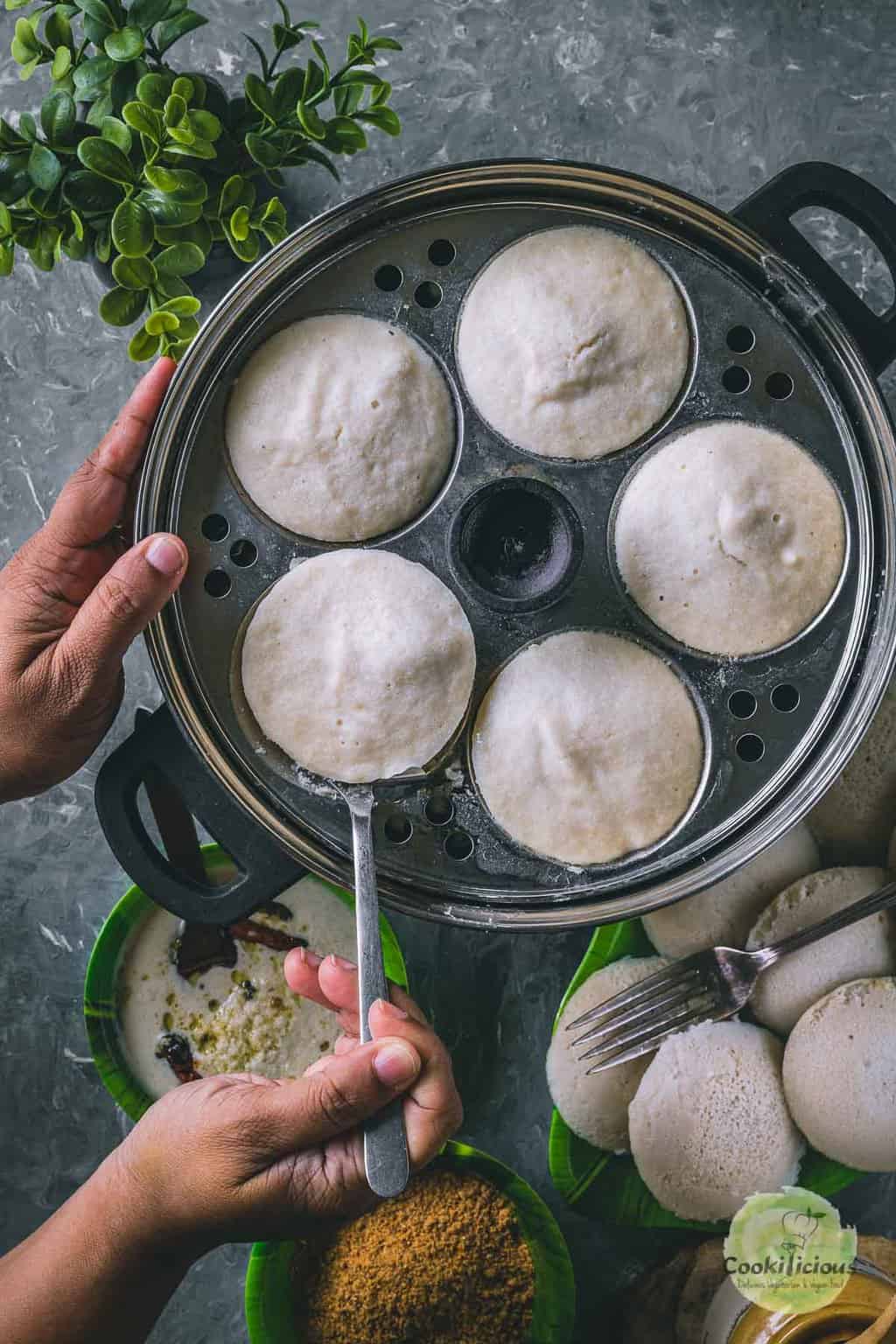 a pair of hands removing idlis from the cooker using a spoon