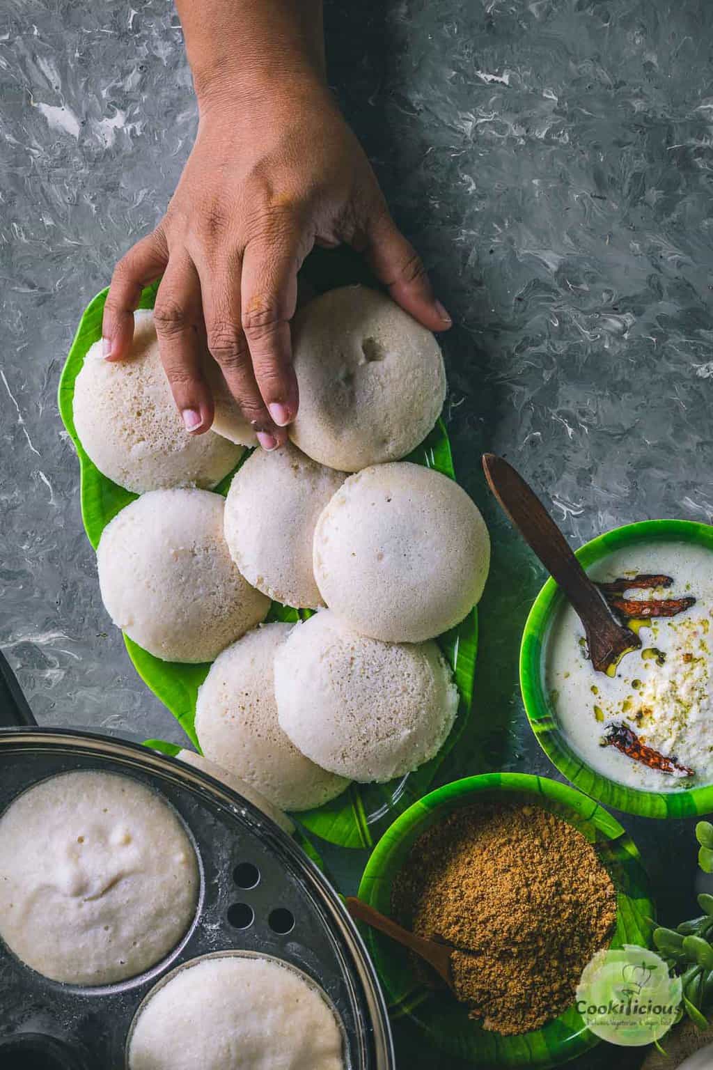 a plate full of white steamed idlis and one hand reaching out to pick one