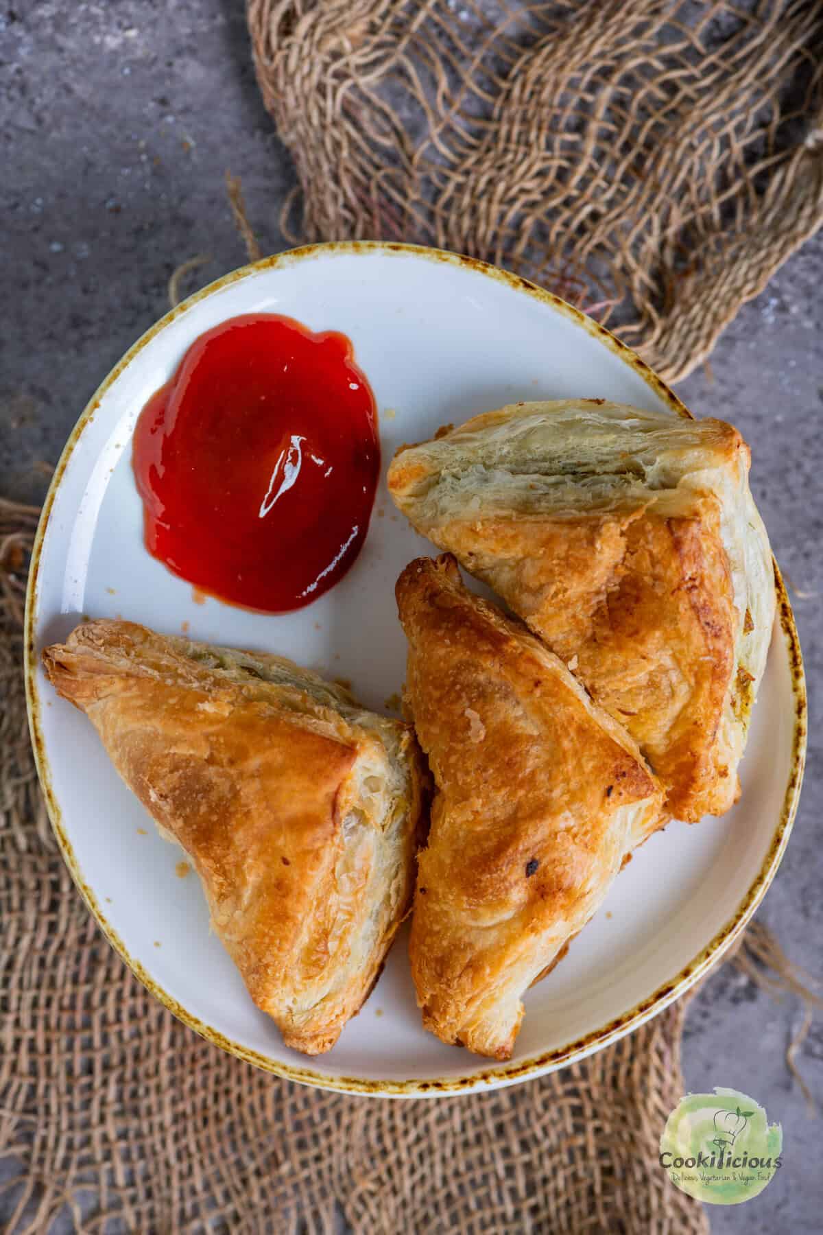3 Crispy puff pastry samosa puffs on a white platter served with tomato ketchup.