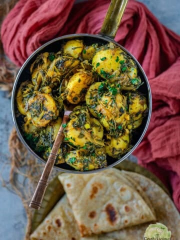 Aloo methi sabzi in a copper pan with a copper spoon, next to a stack of folded phulkas.