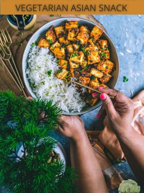 one hand holding the bowl and the other hand digging into the bowl filled with chilli paneer with a fork and text at the top