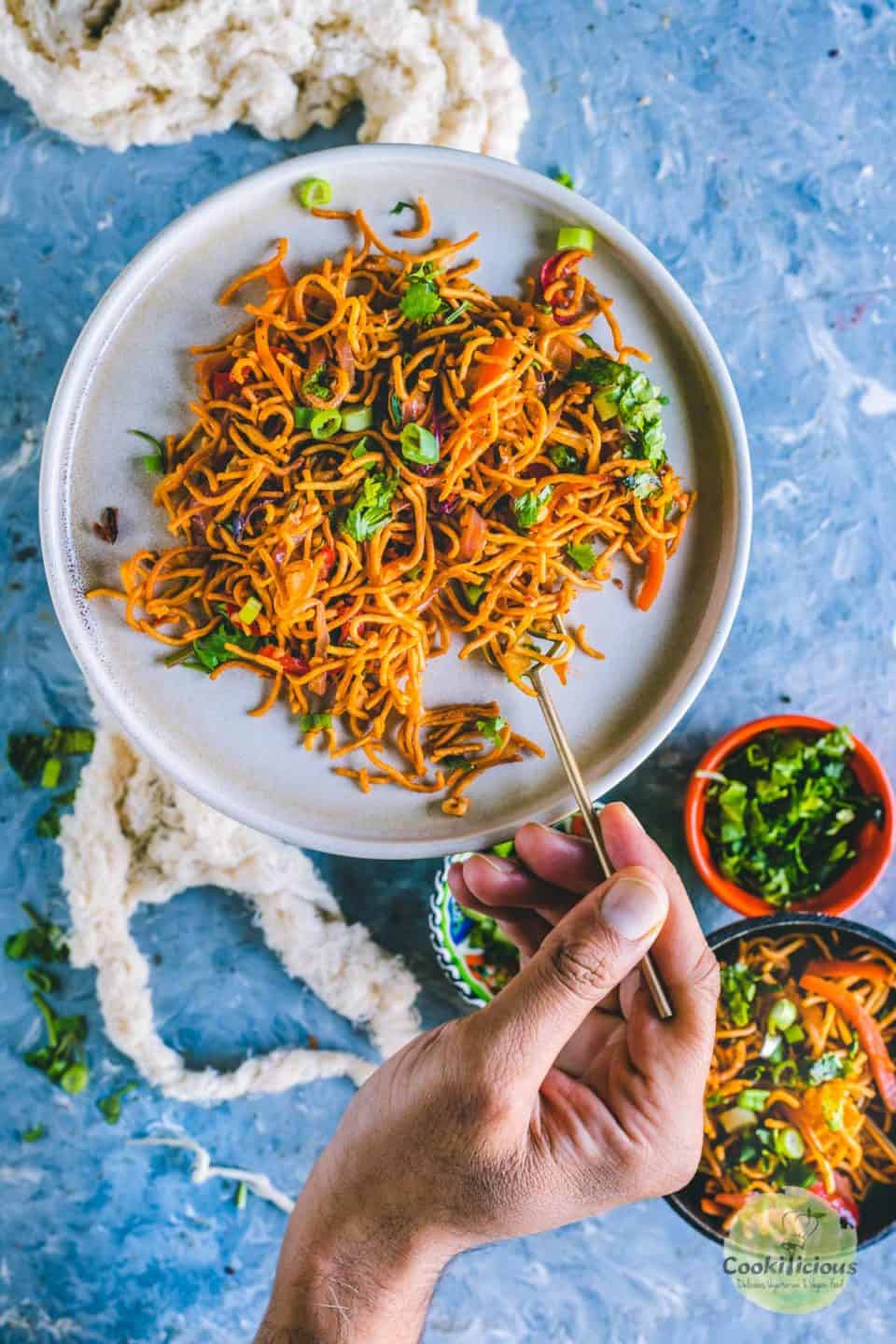 a hand holding a fork and digging into a plate filled with Chinese Bhel 