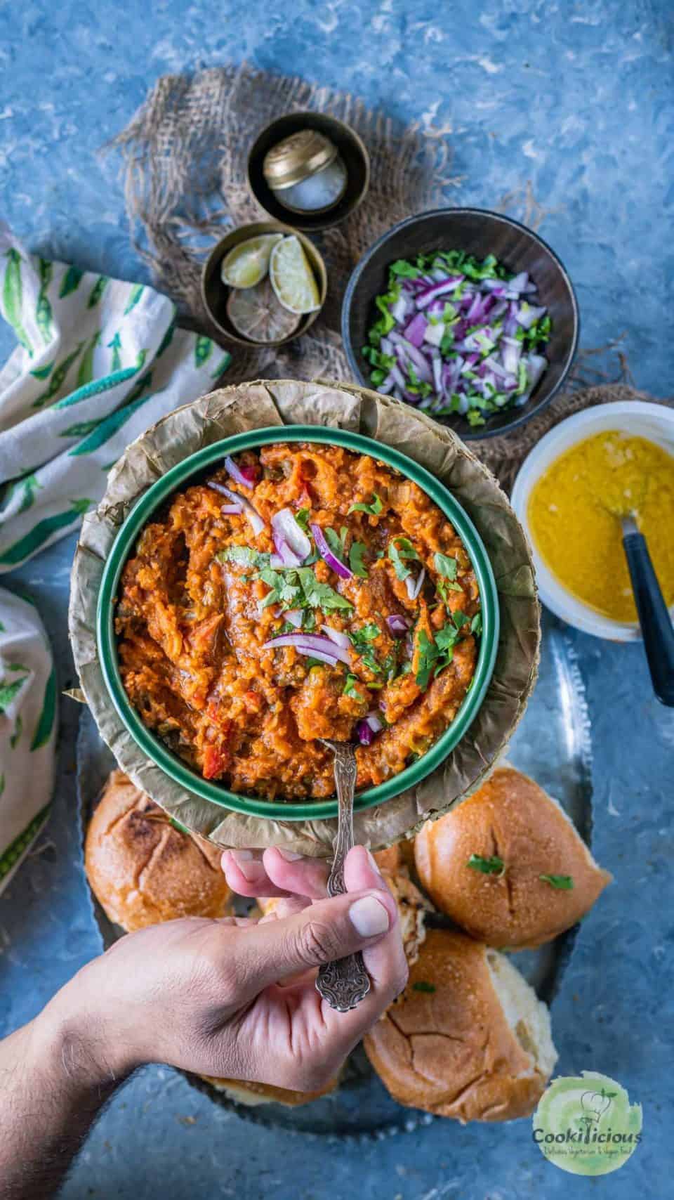 a hand dipping a spoon into a bowl of pav bhaji