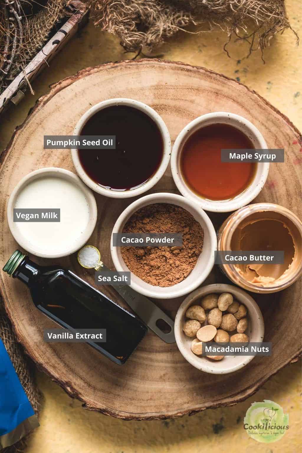 Ingredients for vegan chocolate bars laid out on a wooden board — pumpkin seed oil, maple syrup, vegan milk, cacao powder, cacao butter, sea salt, vanilla extract, and macadamia nuts.