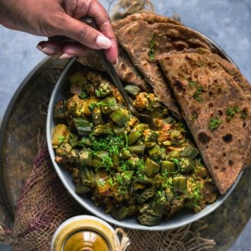 a hand digging into a plate filled with Achari Dahi Bhindi with roti on the side