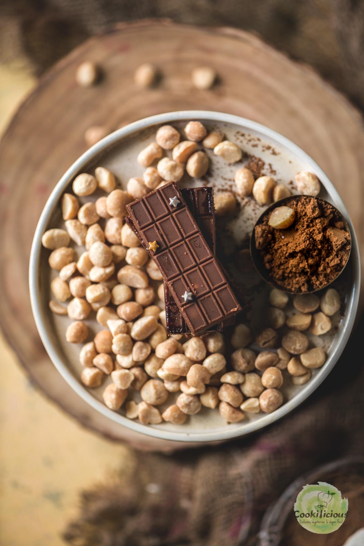 Close-up of mini vegan chocolate bars stacked on a bed of macadamia nuts, with a bowl of cacao powder alongside.