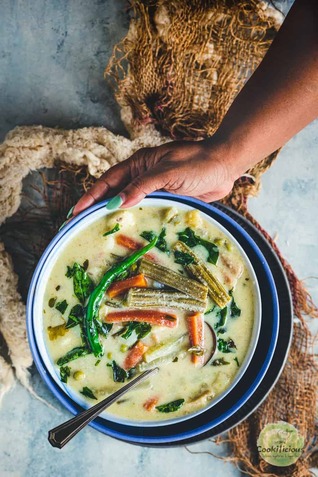 Hand holding a bowl of Tamil-style avial with a spoon, showing the creamy coconut-yogurt curry with mixed vegetables.