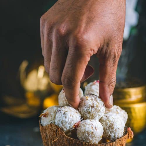 a hand picking up one coconut ladoo from a bowl