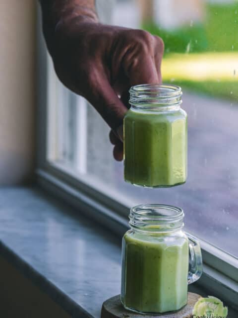 one hand picking up one glass of cold cucumber soup while another one sits next to it