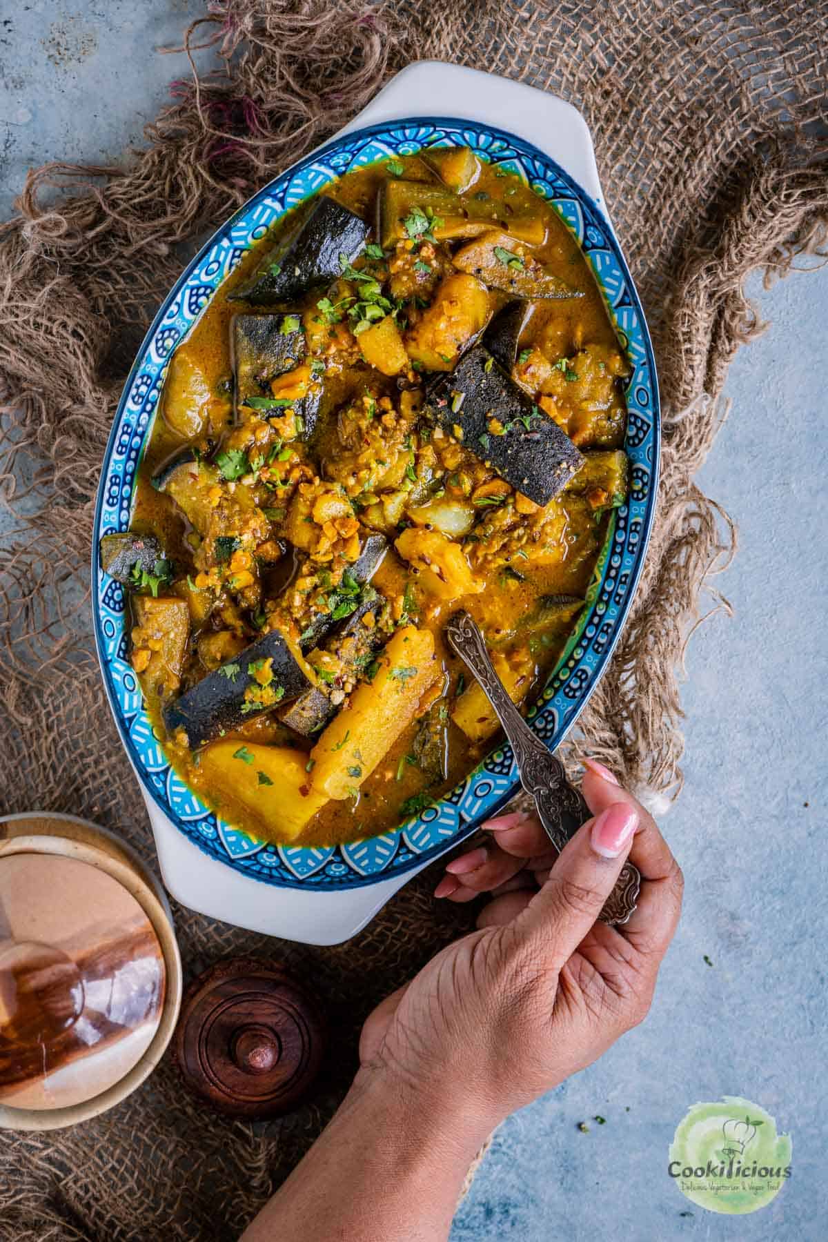 a hand digging into a bowl of Aloo Baingan with a spoon