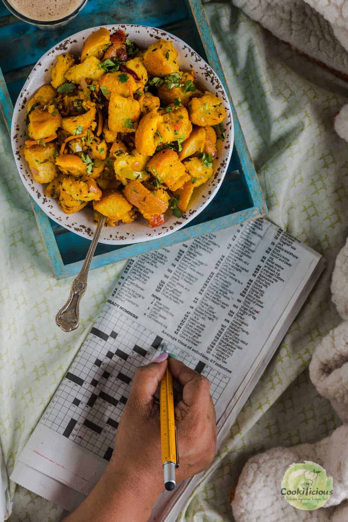 a bowl of Bread Upma placed next to a person solving a crossword on a newspaper