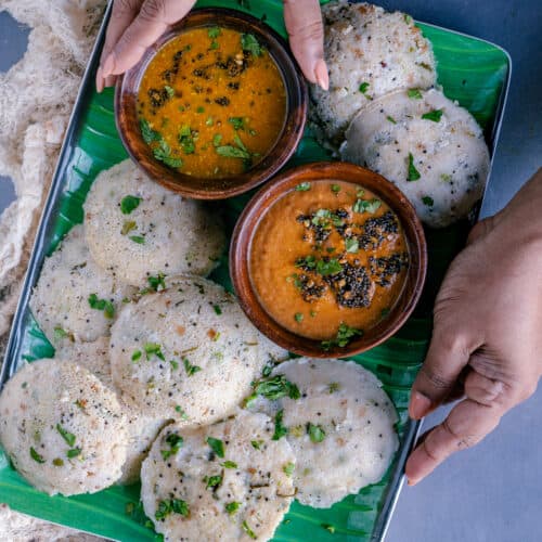 a set of hands holding a tray of rava idli with bowls of sambhar and chutney in it.