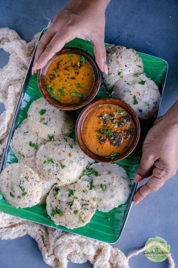 a set of hands holding a tray of rava idli with bowls of sambhar and chutney in it.