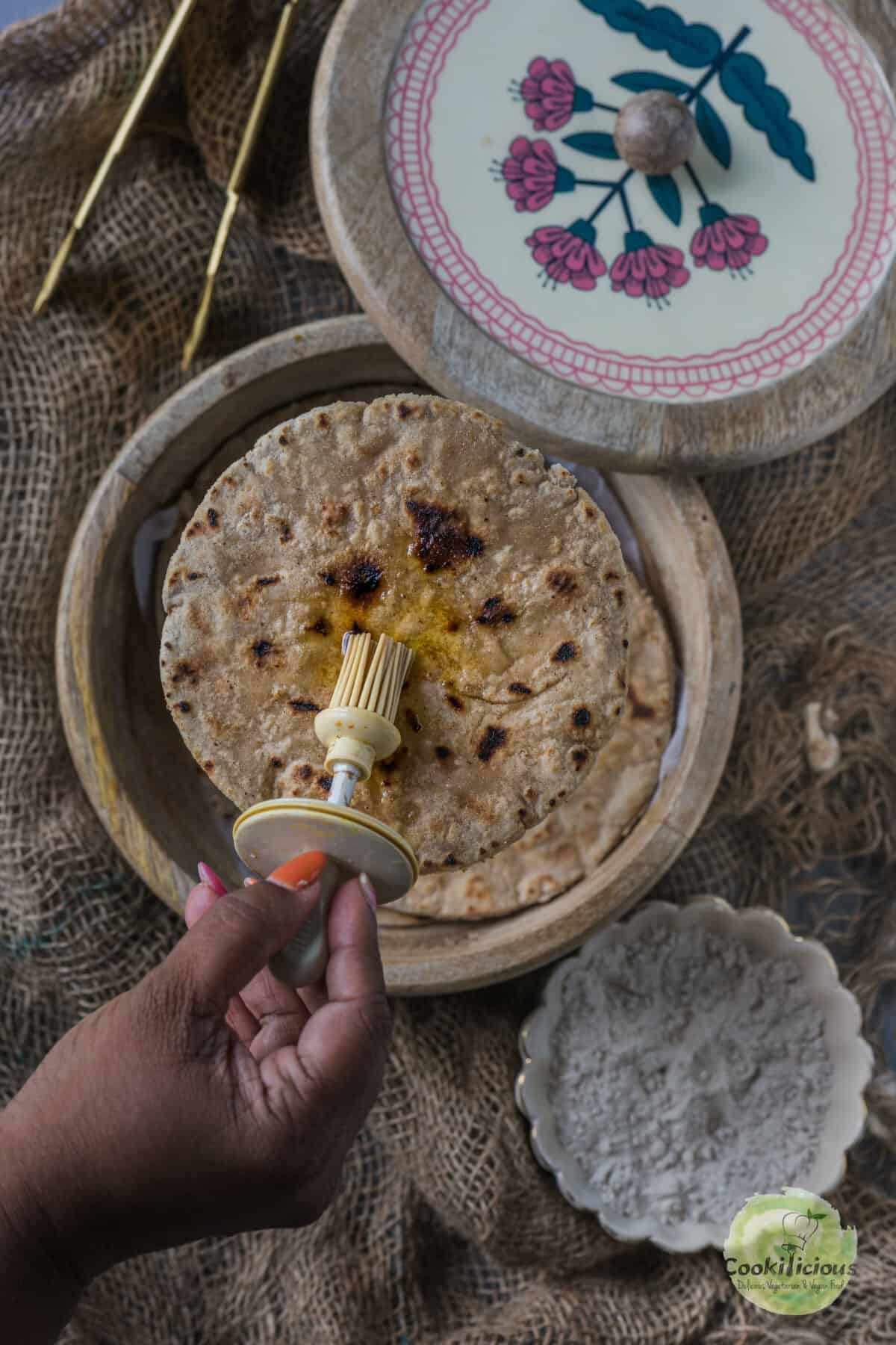 a hand applying ghee on roti that's placed in a box.