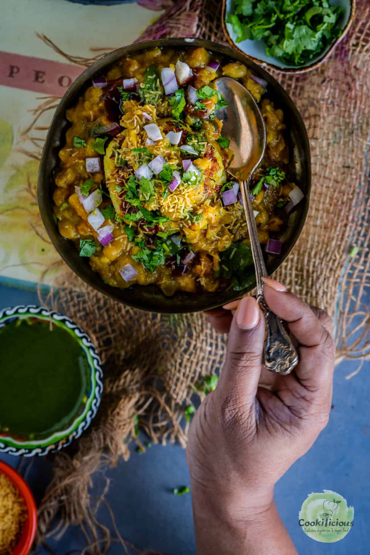 a hand digging into a plate of 2 bowls of Ragda Pattice, holding a spoon. 