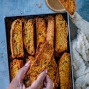 A hand picking up a savory jalapeño cheddar cornbread biscotti from a tray full of eggless biscotti, with one slice resting on a glass of coffee.