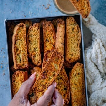 A hand picking up a savory jalapeño cheddar cornbread biscotti from a tray full of eggless biscotti, with one slice resting on a glass of coffee.