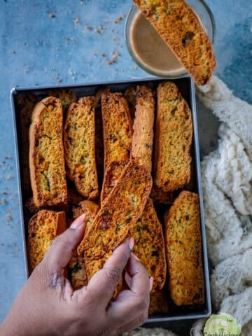 A hand picking up a savory jalapeño cheddar cornbread biscotti from a tray full of eggless biscotti, with one slice resting on a glass of coffee.