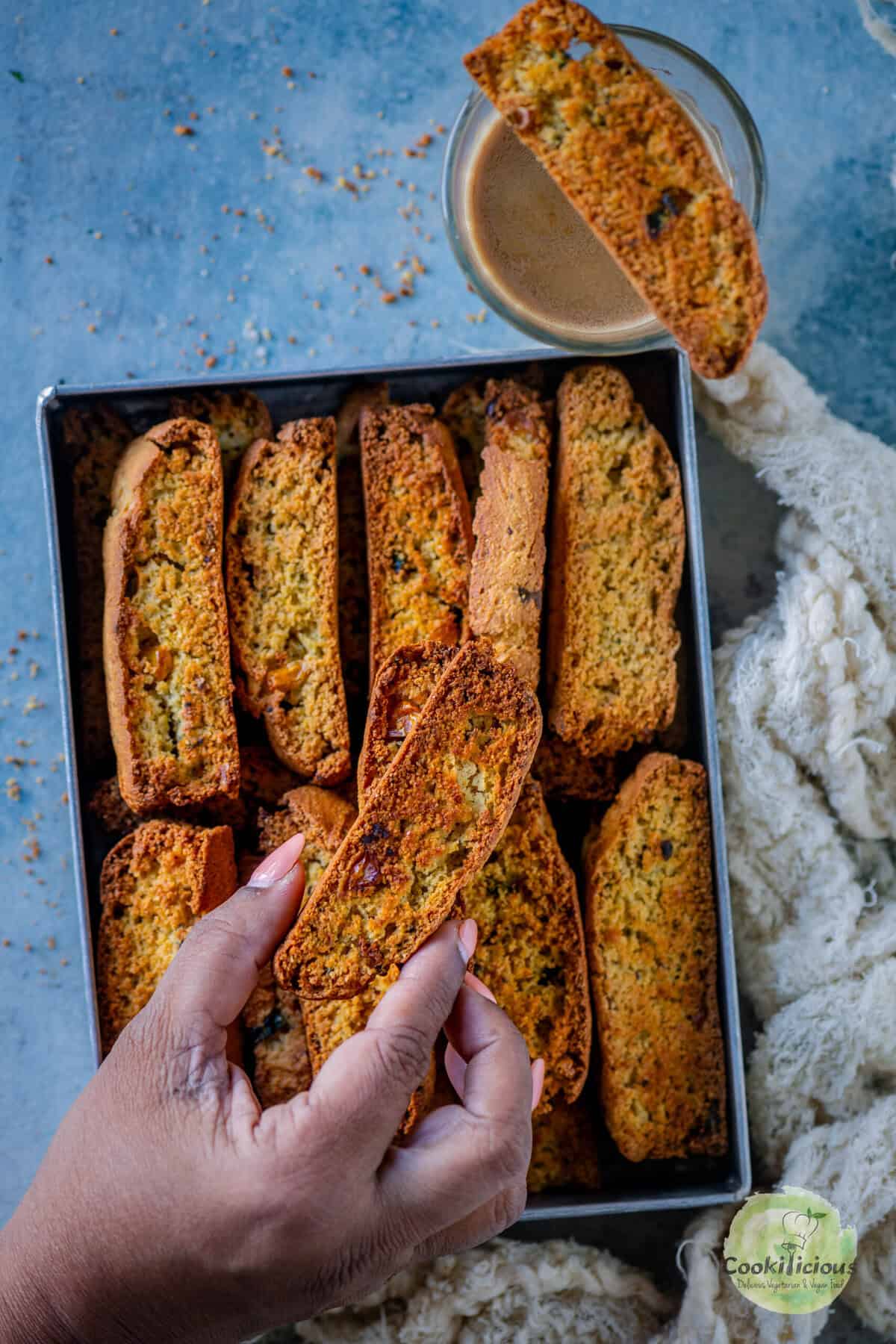 A hand picking up a savory jalapeño cheddar cornbread biscotti from a tray full of eggless biscotti, with one slice resting on a glass of coffee.