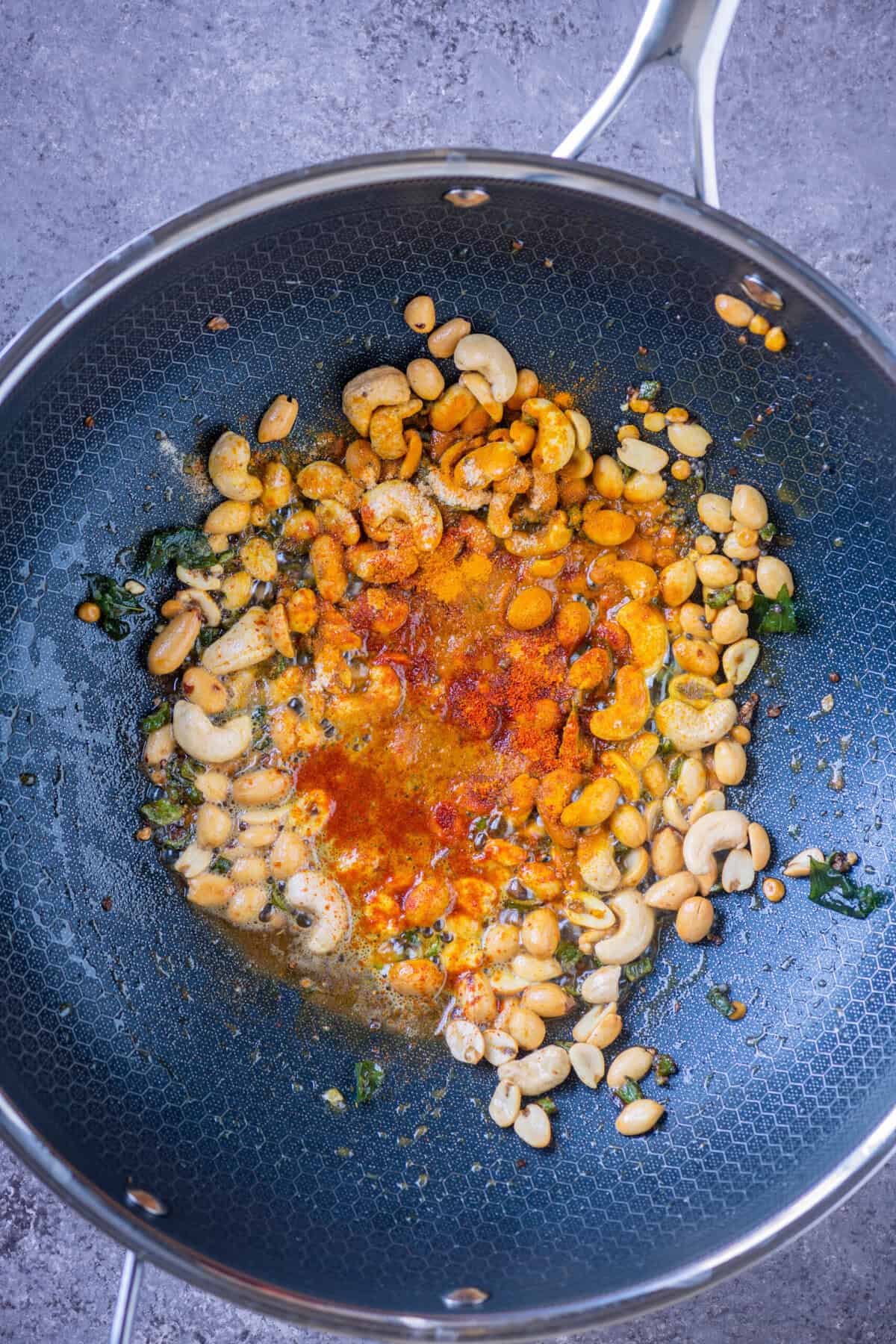 Turmeric, red chilli powder, and asafoetida sprinkled over fried peanuts and cashews in a wok.