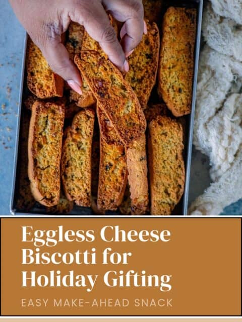 A hand picking up an eggless cheese biscotti from a tray of savory biscotti, with text reading “Eggless Cheese Biscotti for Holiday Gifting – Easy Make-Ahead Snack.
