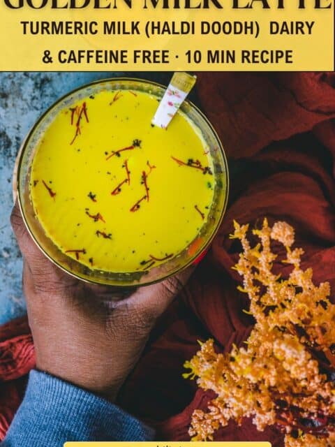 Overhead view of a hand holding a turmeric milk (haldi doodh) latte topped with saffron strands and black pepper.