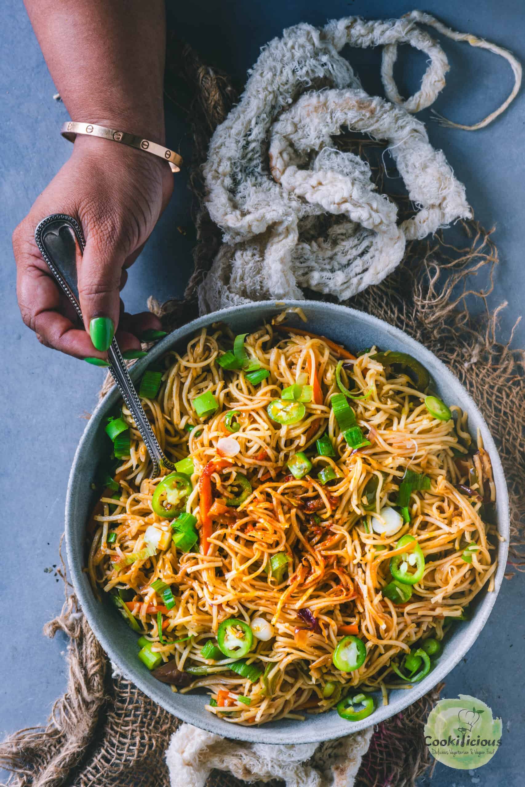 A hand holding a fork and digging into a bowl of Veg Hakka Noodles.