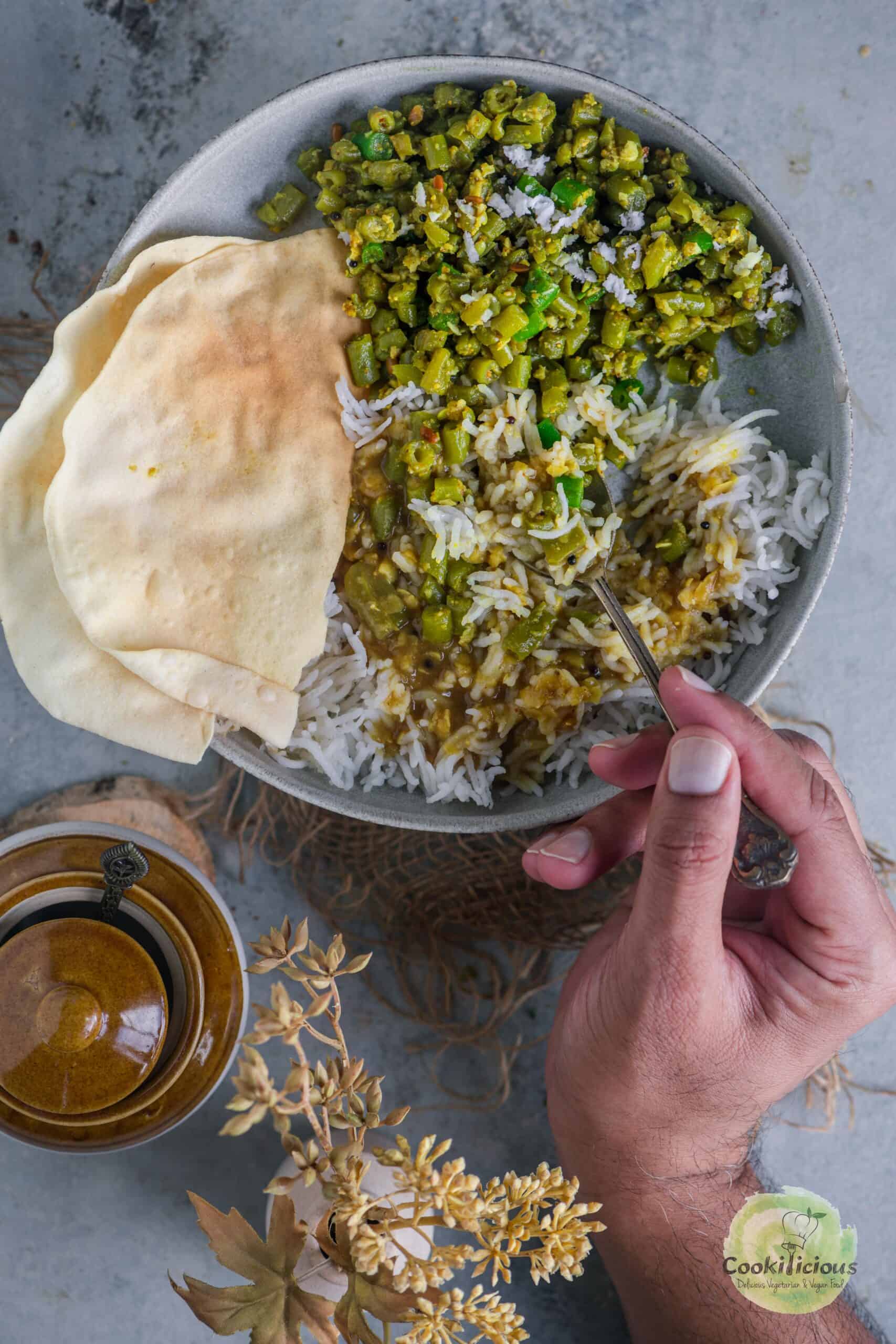 A hand digging into a bowl of Beans Thoran that's served with rasam, rice and papad.