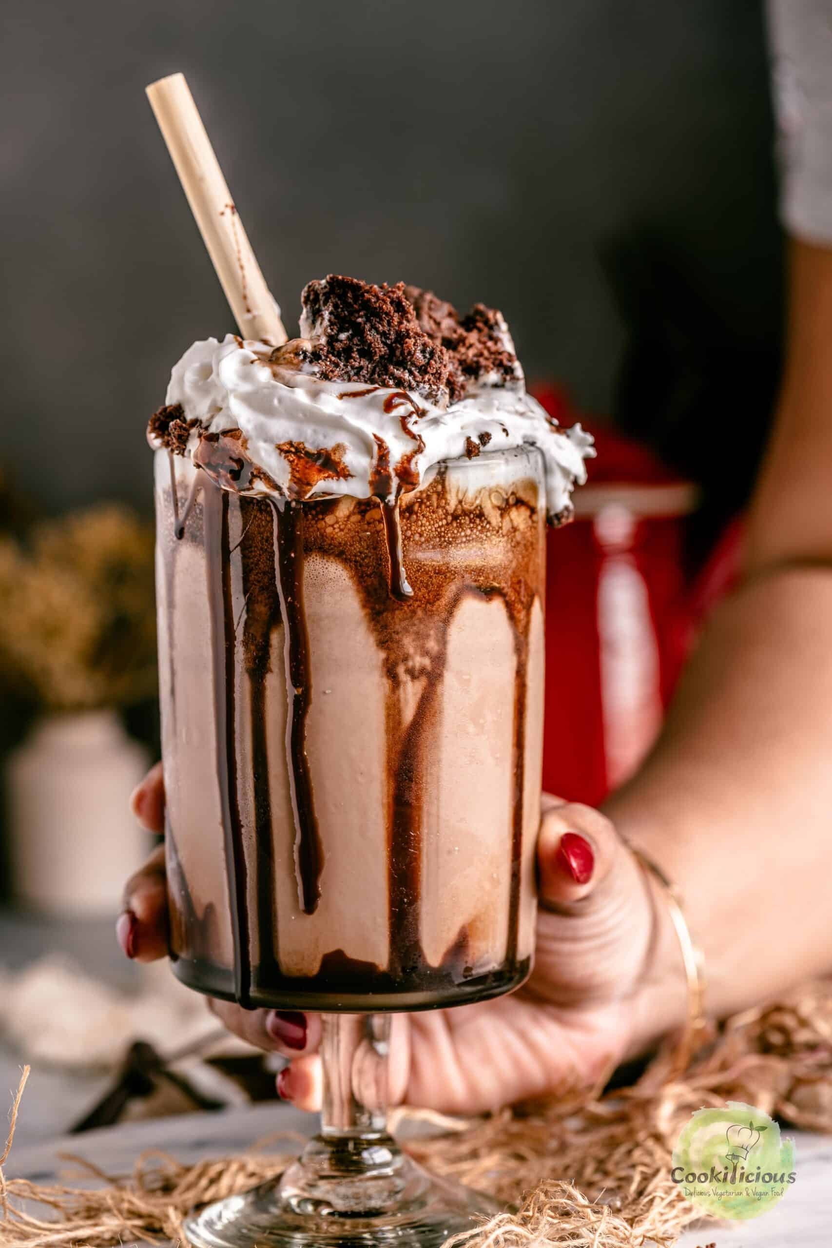 A tall glass of homemade brownie milkshake, garnished with whipped cream, a fudgy brownie wedge and a straw, as a hand lifts it from the table.