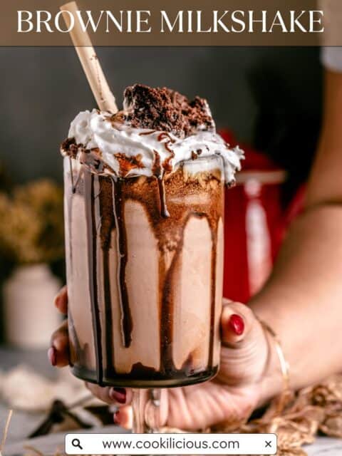 A tall glass of homemade brownie milkshake, garnished with whipped cream, a fudgy brownie wedge and a straw, as a hand lifts it from the table and text at the top and bottom.