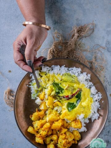 Mor Kuzhambu served with rice and potato Fry in a bowl and a hand digging into it with a spoon.