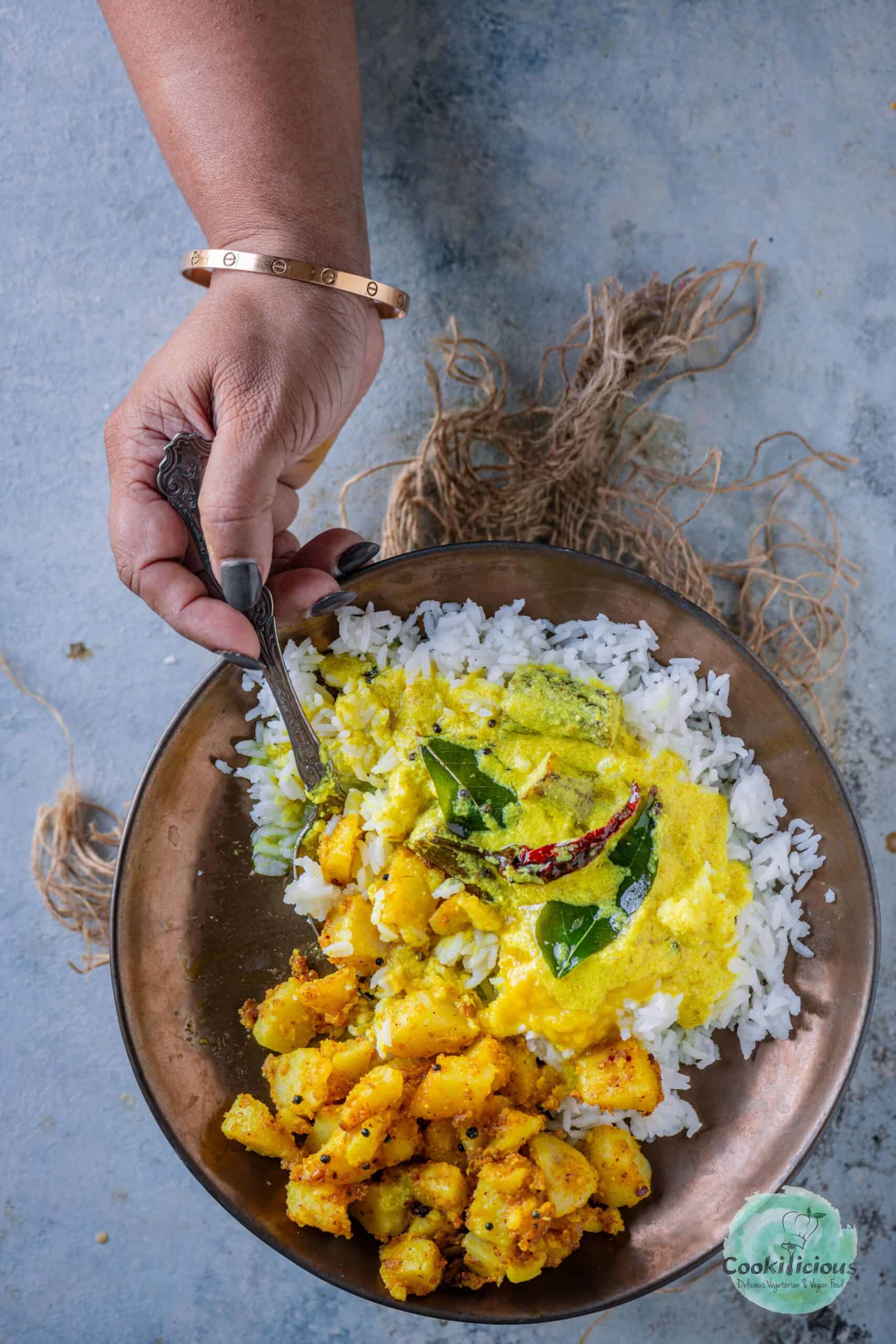 Mor Kuzhambu served with rice and potato Fry in a bowl and a hand digging into it with a spoon.