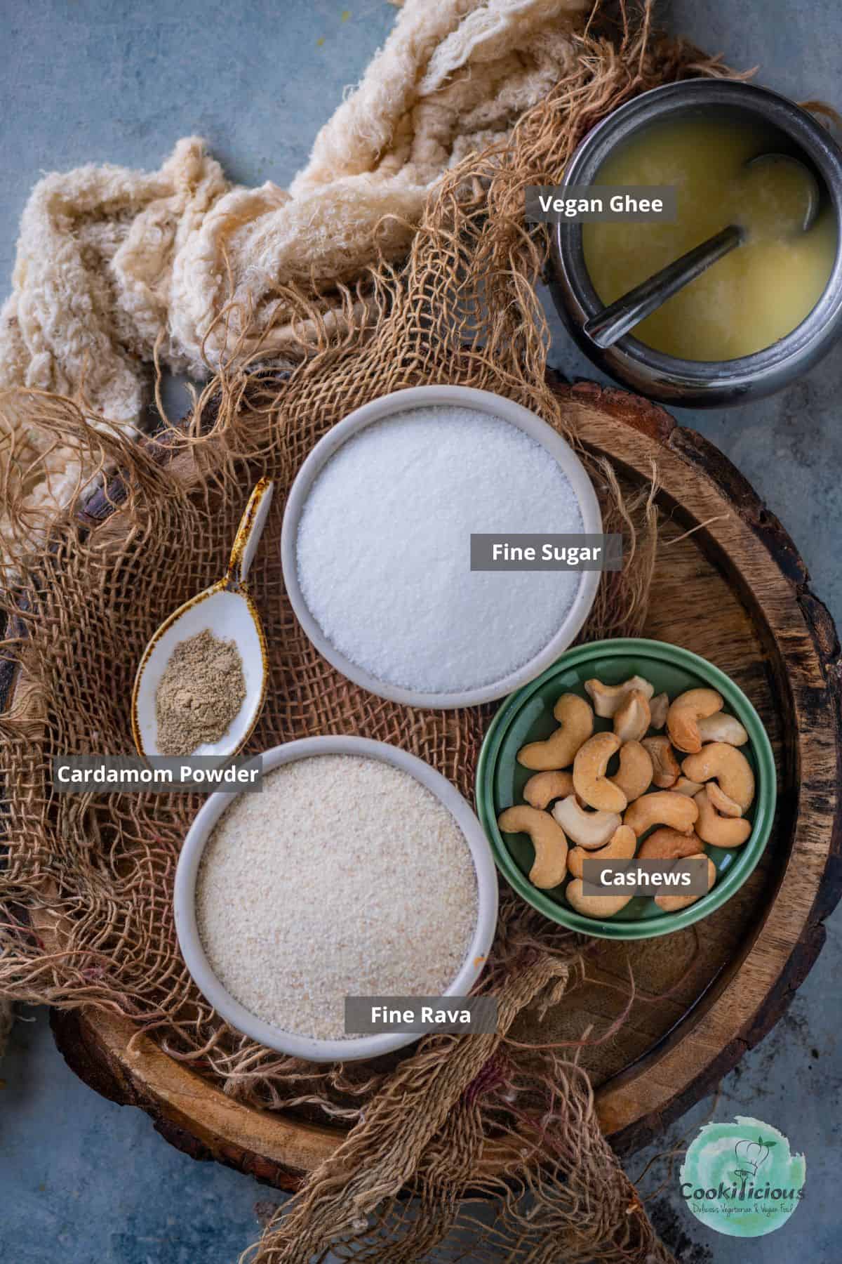 Ingredients for vegan Rava Laddu: semolina, sugar, cashews, cardamom, and vegan ghee placed on a table with labels on them.