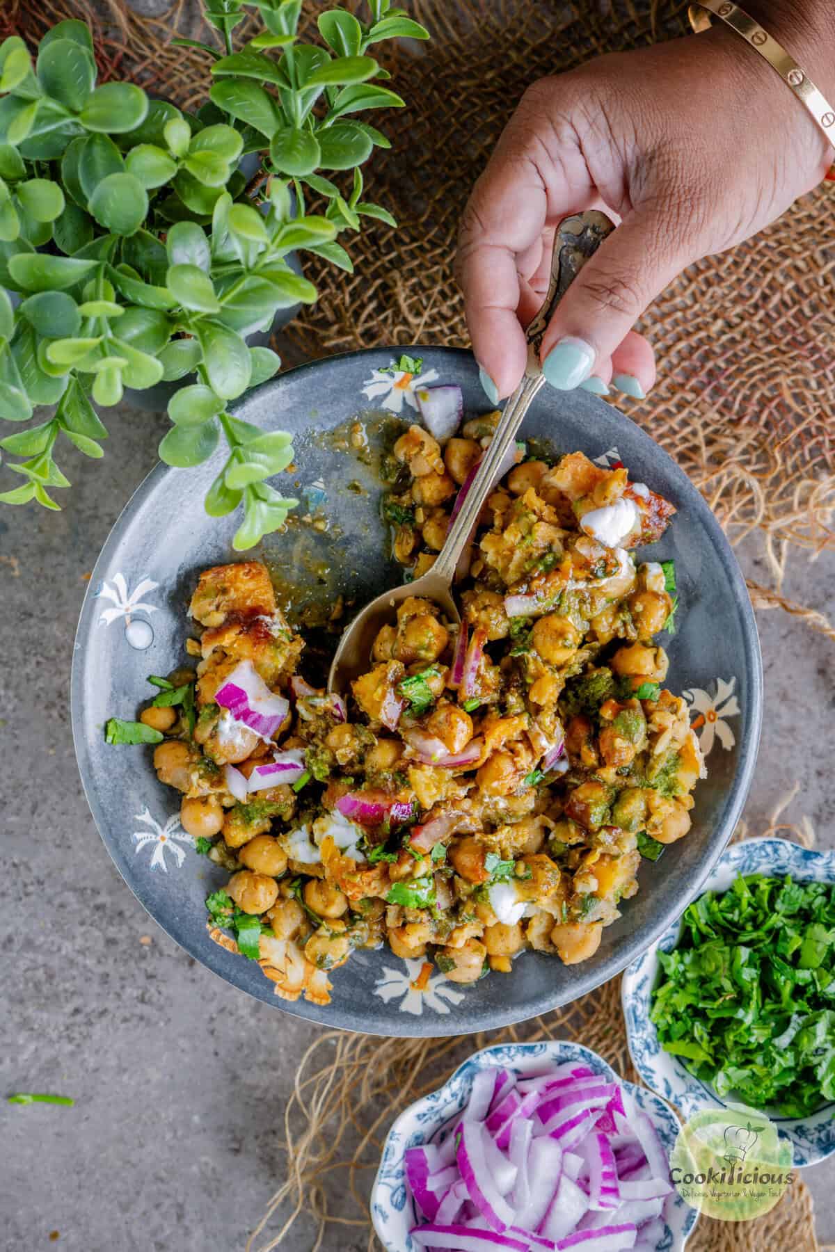 Close-up of street-style samosa chaat with chickpea curry, meetha chutney, green chutney, yogurt drizzle, onions, cilantro, and sev topping.