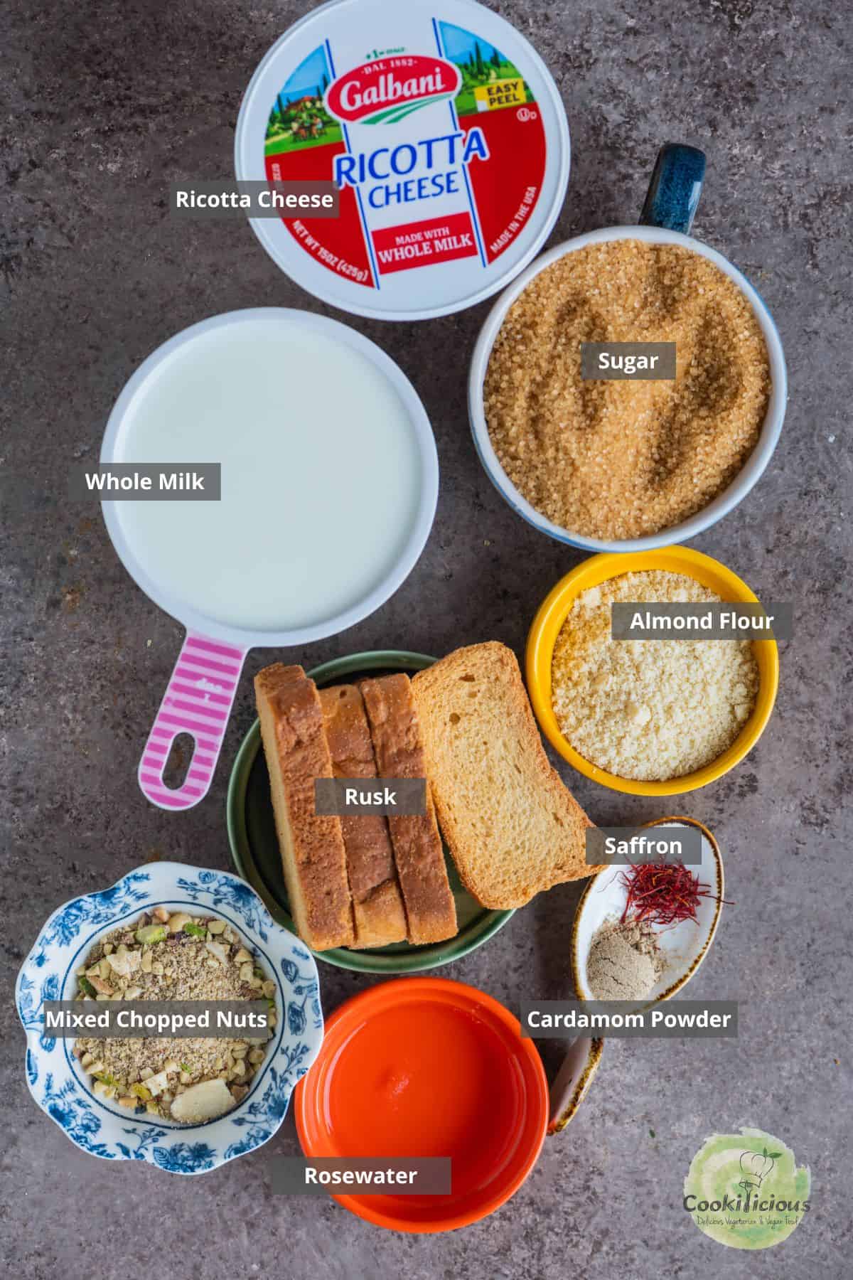 Ingredients for Rabdi Cheesecake — ricotta cheese, rusk, saffron, cardamom, and chopped pistachios arranged on a countertop with labels on them.